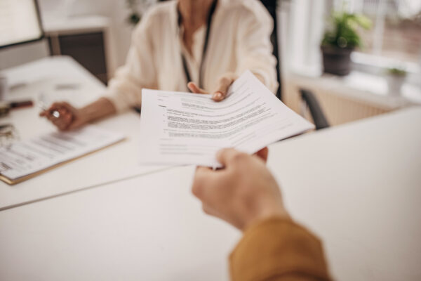 Two people, mature businesswoman sitting at her desk in office, giving documents to young colleague. Image by South_agency, istockphoto.com