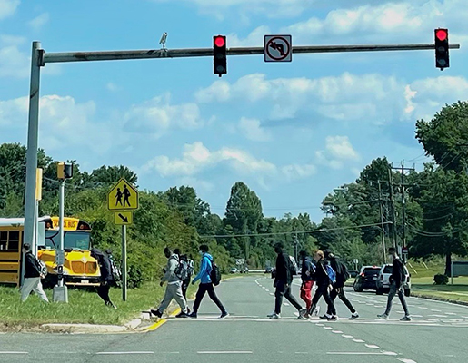 A group of students wearing backpacks and casual clothes cross at a crosswalk in front of Largo High School.